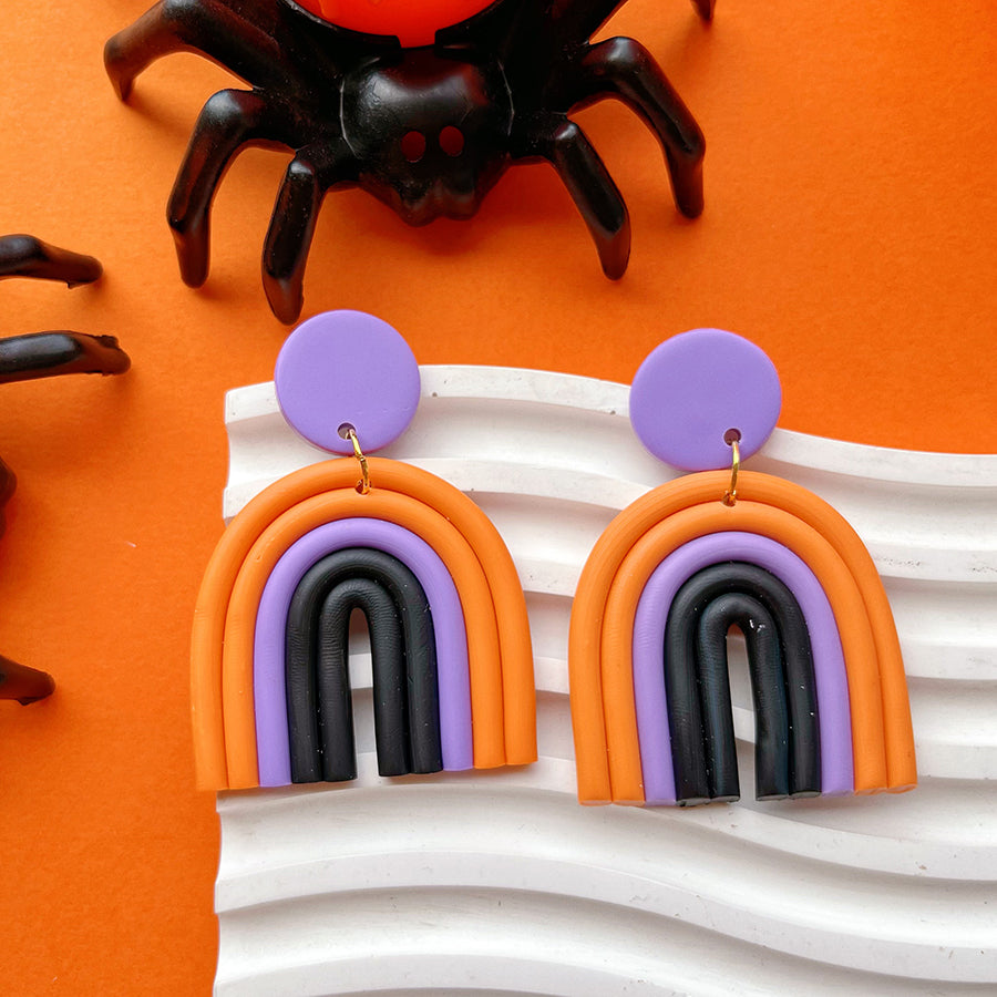 Woman wearing a black headband with colorful designs, purple earrings, and a gold necklace against an orange background.