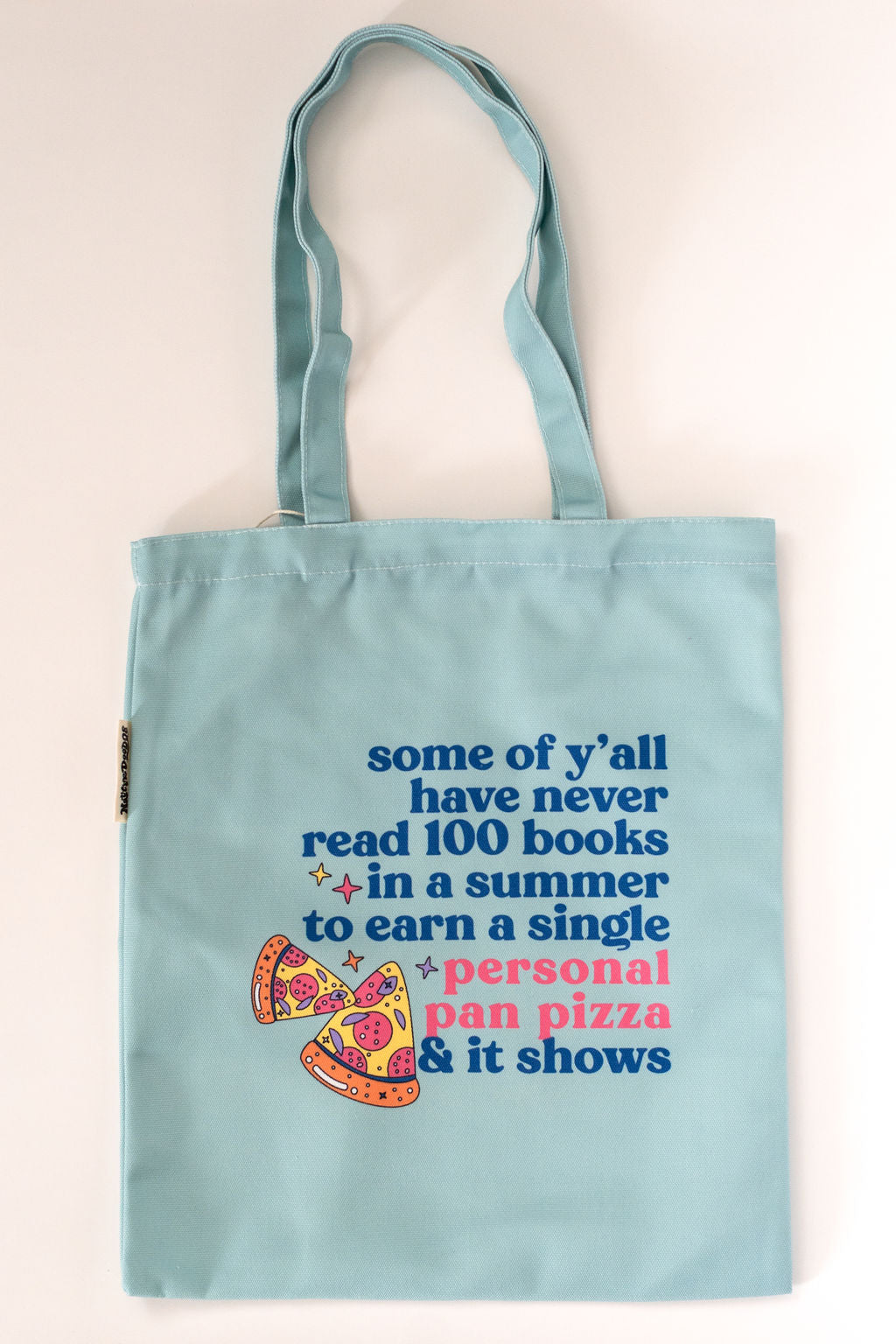 Light blue tote bag with colorful text and pizza graphic on a beige background