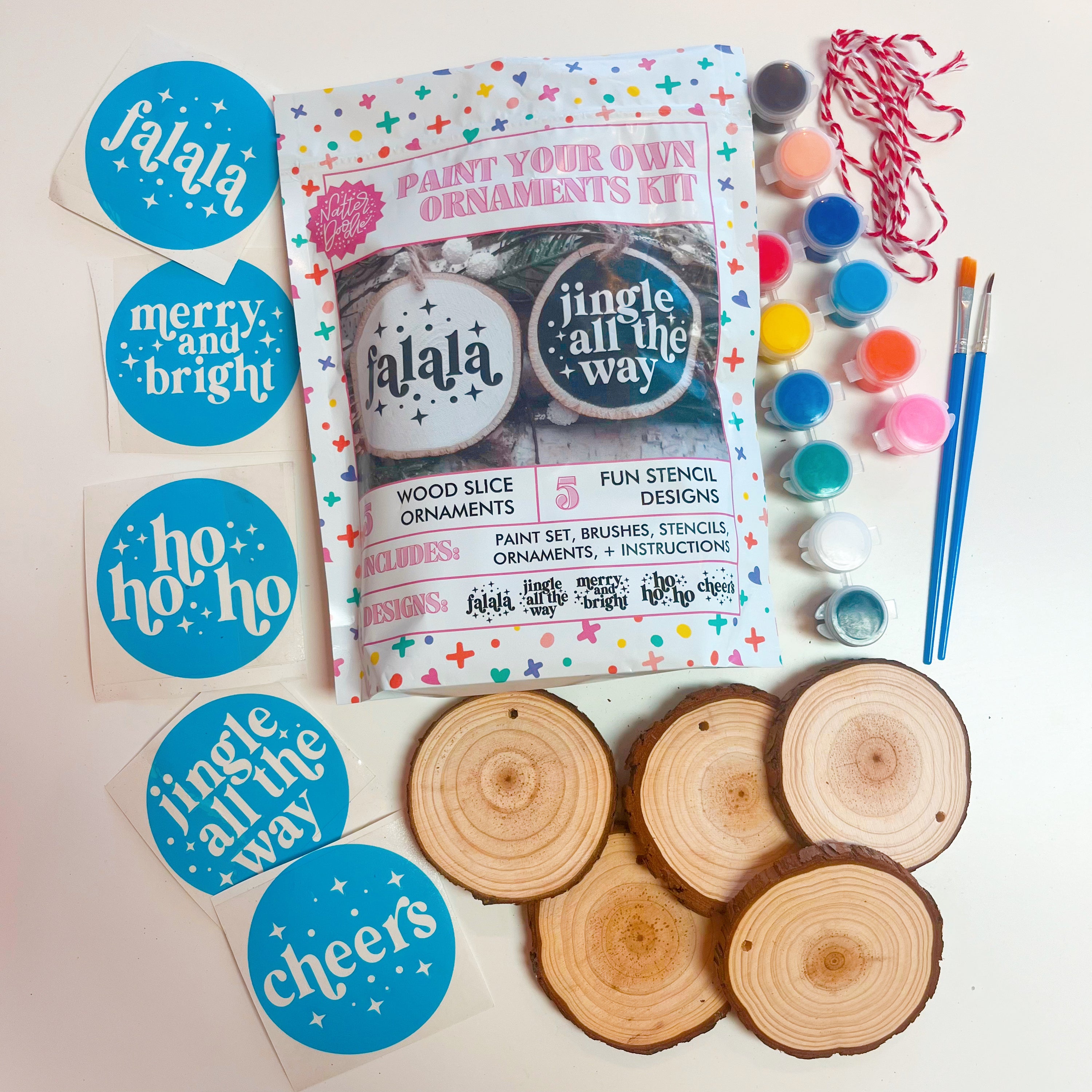 A kit to paint your own holiday ornaments, including 5 sliced birch wood ornaments with bark, a set of hand lettered design stencils, a paint set with 12 colors, and two paint brushes, with the packaging showing examples of the finished ornaments.