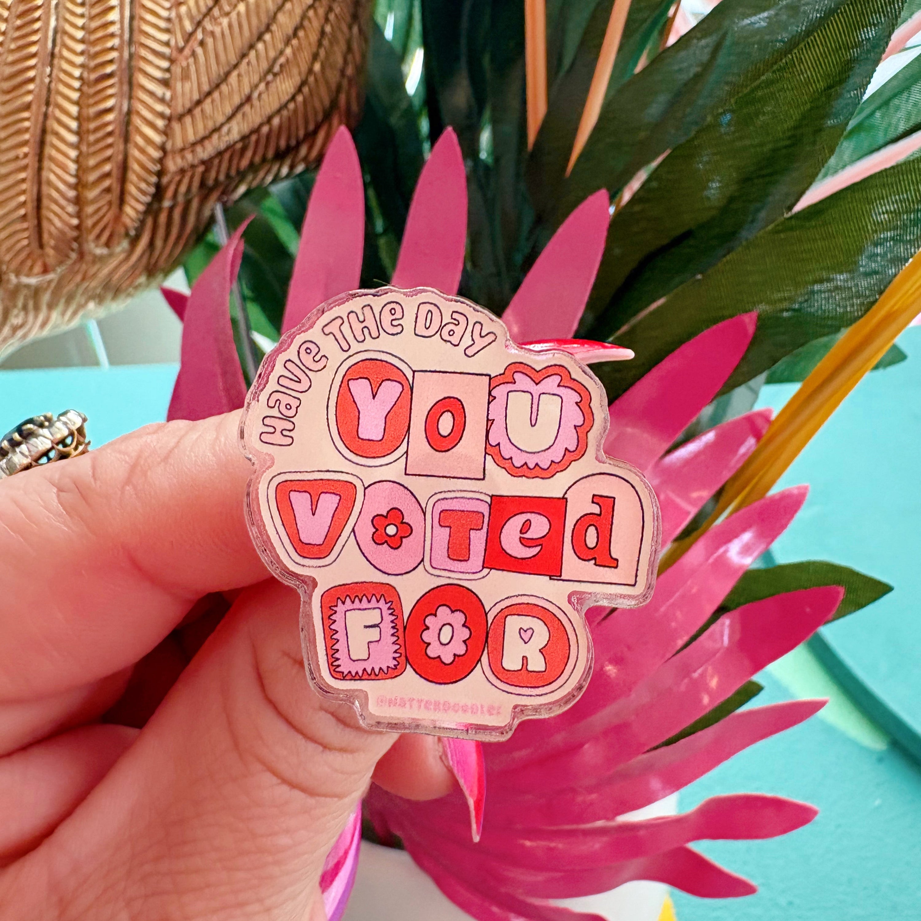 An acrylic pin with the text 'Have the Day You Voted For' printed in red and transparent colors, held in a hand with a colorful background.