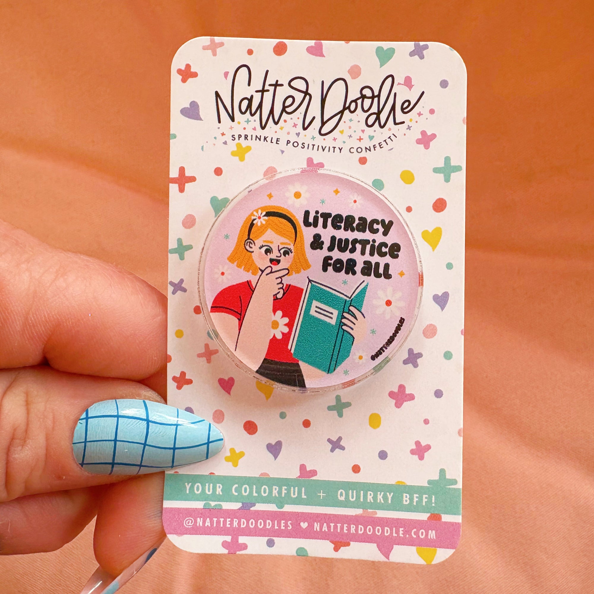 A pin with an illustration of a cartoon book character and the text 'Literacy & Justice For All' on a pink background, held by a hand with a blue grid-patterned nail polish.
