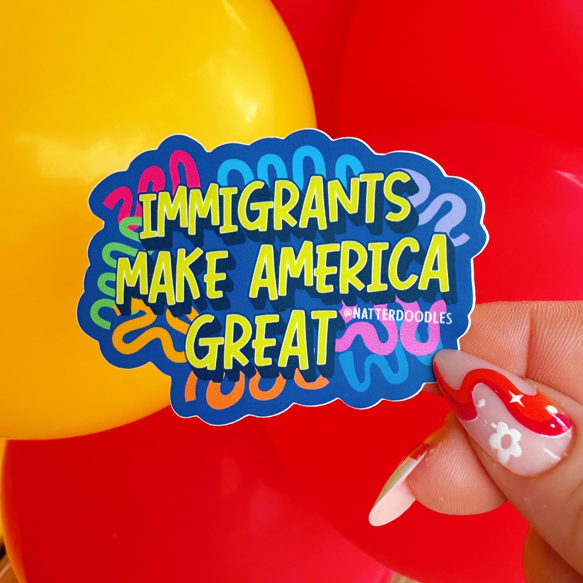 Colorful sticker with 'Immigrants Make America Great' text held in front of red and yellow balloons.