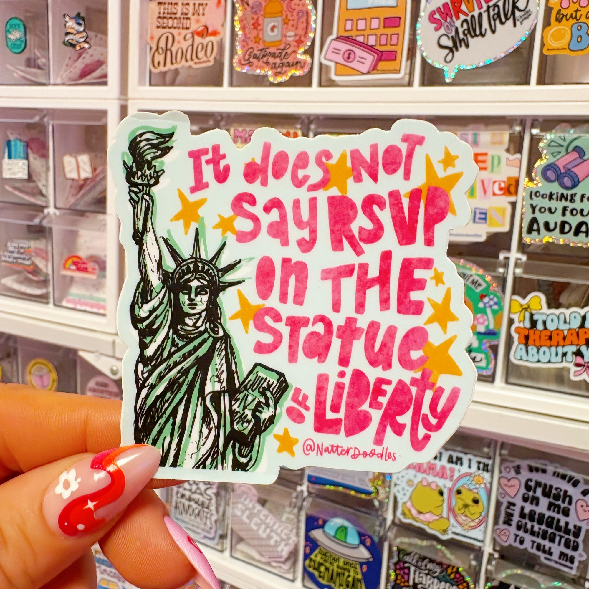 Sticker with text 'It does not say RSVP on THE STATUE OF LIBERTY' held by a hand in front of a display of other stickers.