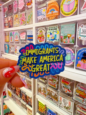 Sticker with 'Immigrants Make America Great' text held in front of a shelf with various small packages.