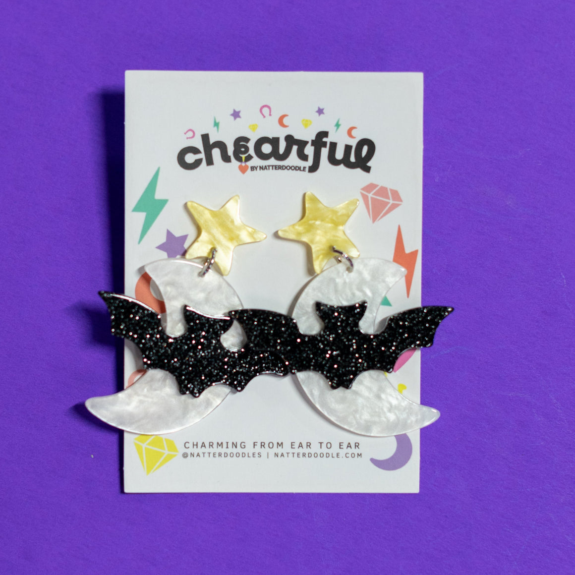 Pair of black bat-shaped earrings on a 'cheerful' branded card against a purple background