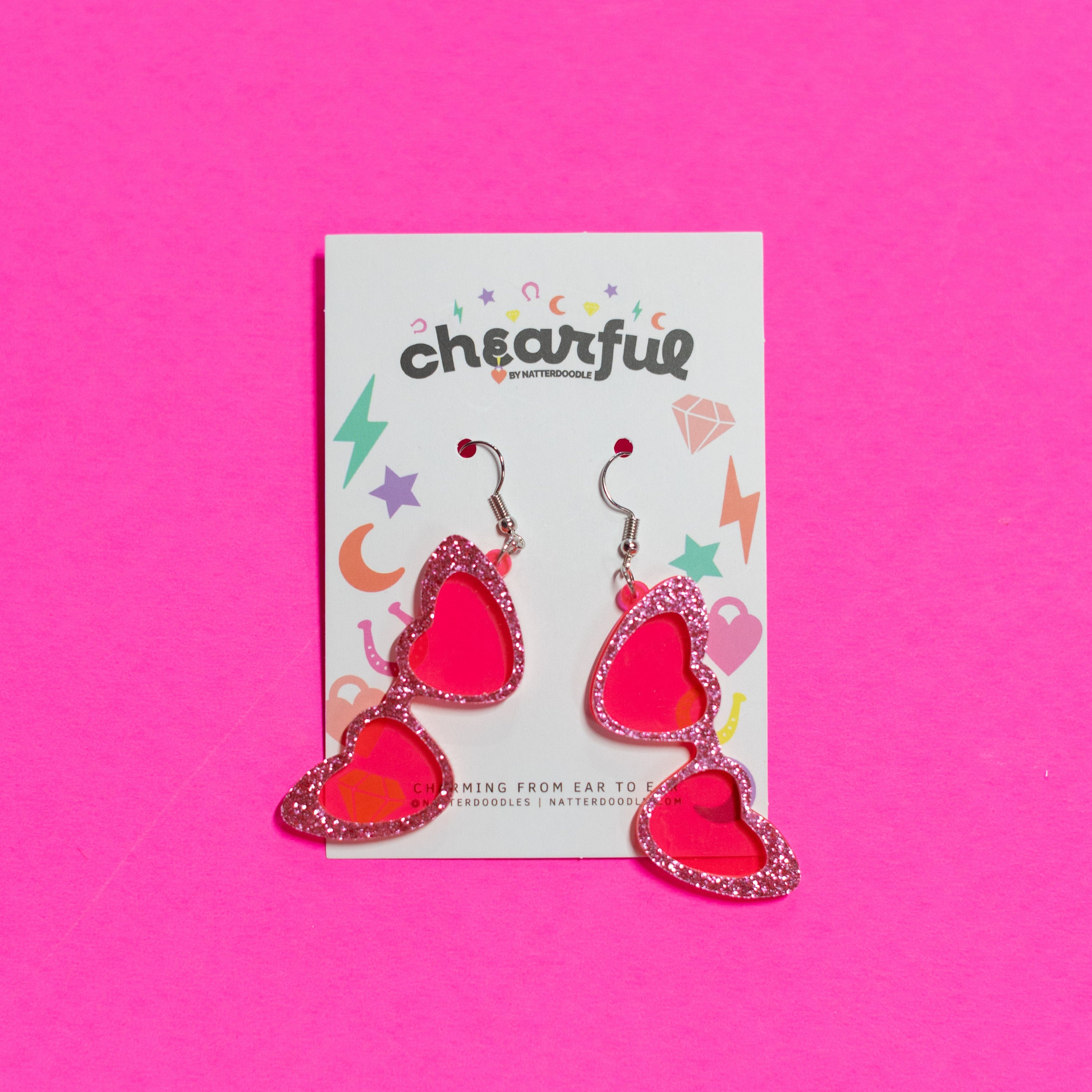 Heart-shaped earrings on a 'cheerful' branded card against a pink background