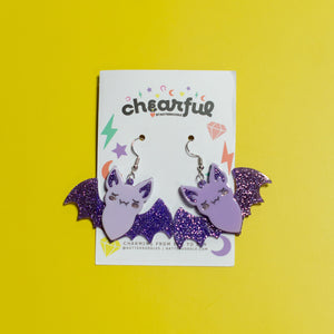 Bat-shaped earrings on a 'cheerful' branded card against a yellow background