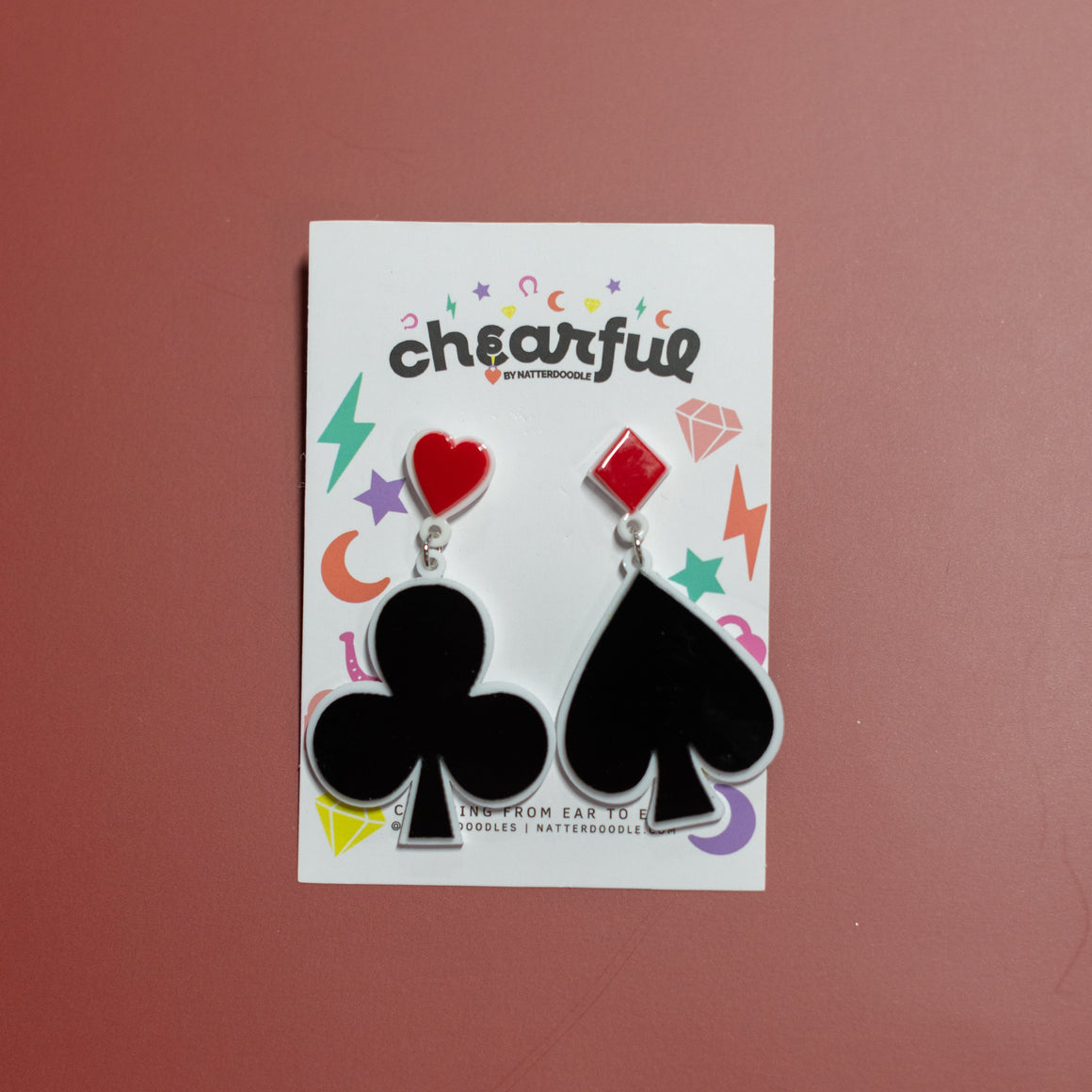 Pair of earrings with club and spade symbols on a 'cheerful' branded card against a red background