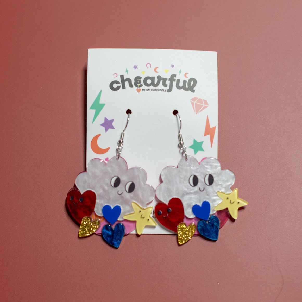 Pair of cloud-shaped earrings with colorful accessories on a 'cheerful' branded card against a pink background.