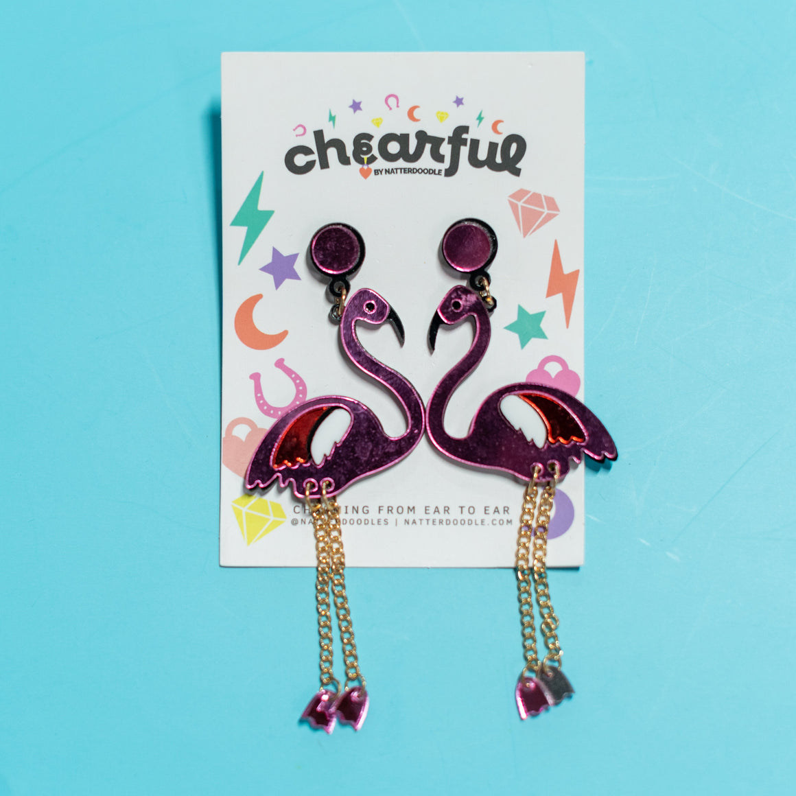 Flamingo-shaped earrings on a 'cheerful' branded card against a blue background