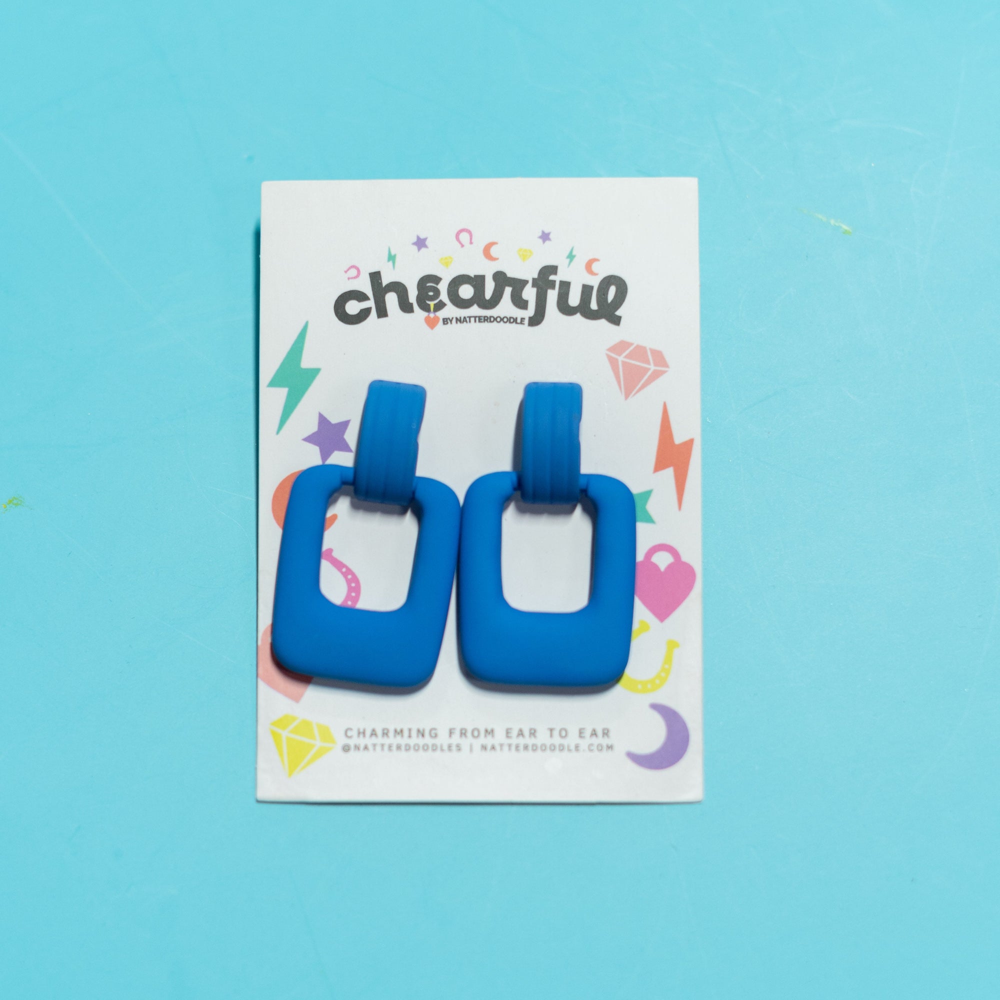 Blue square-shaped earrings on a 'cheearful' branded card against a blue background