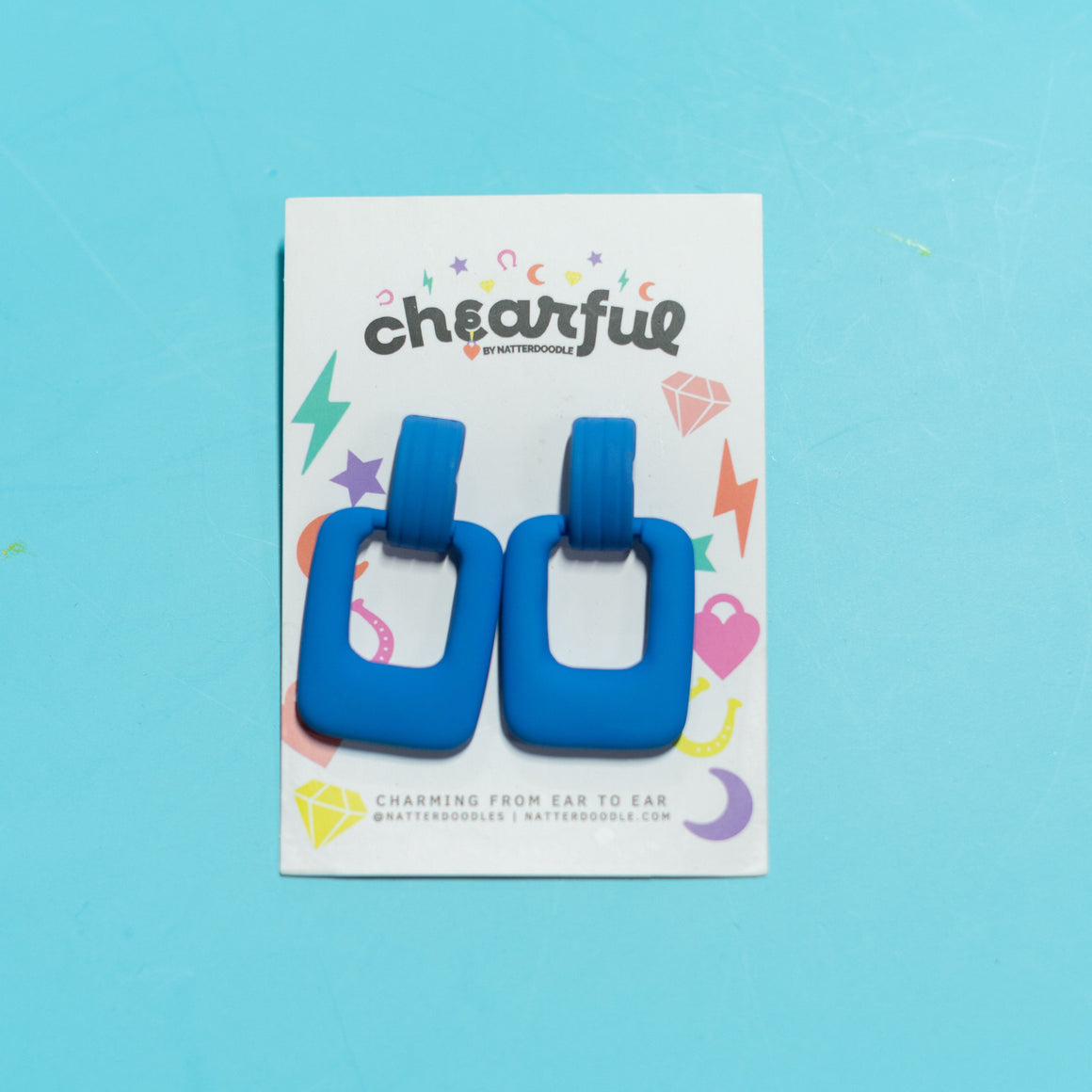 Blue square-shaped earrings on a 'cheearful' branded card against a blue background