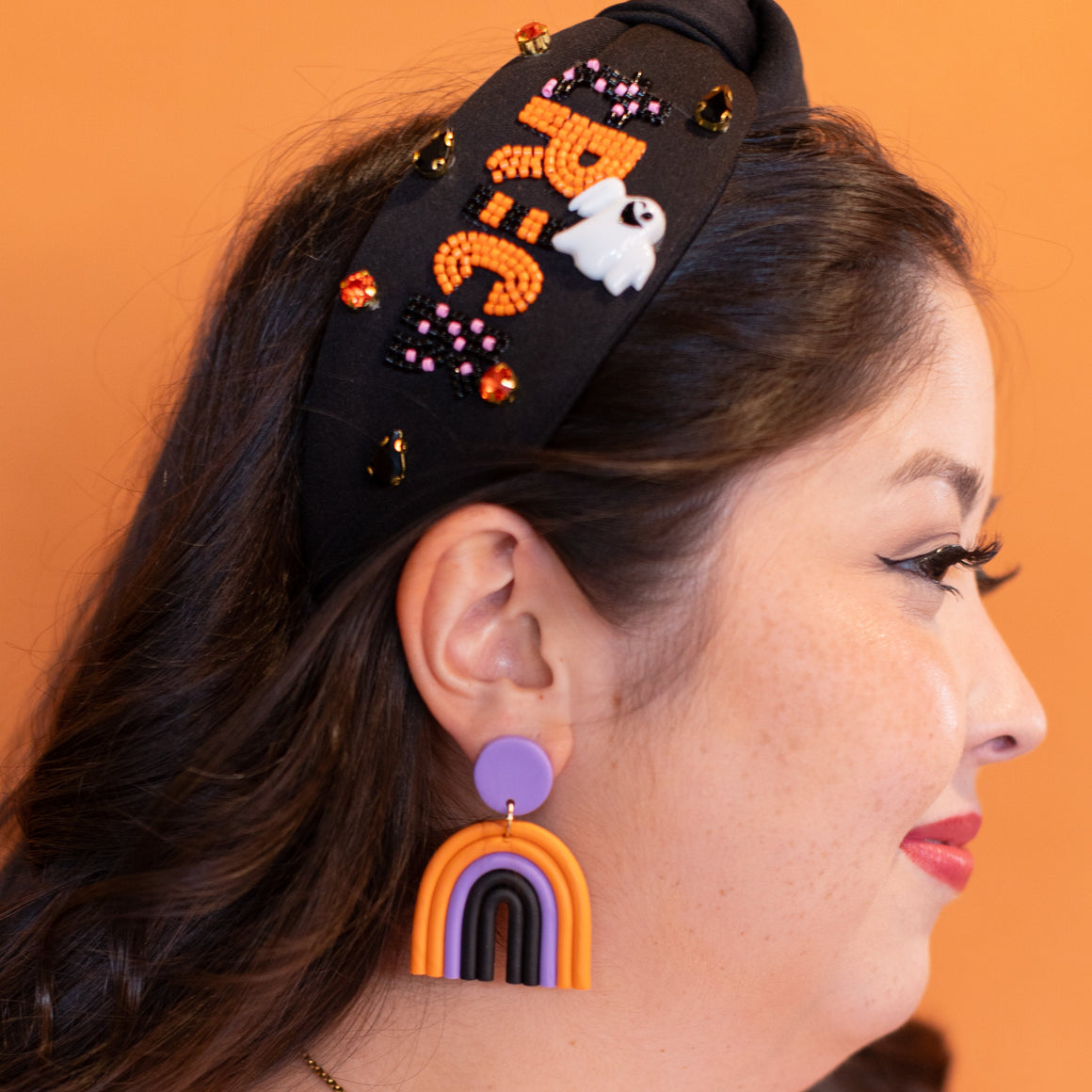 Woman wearing a black headband with colorful designs, purple earrings, and a gold necklace against an orange background.