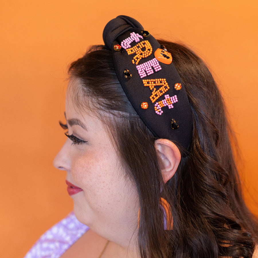 Woman wearing a black headband with colorful designs, purple earrings, and a gold necklace against an orange background.