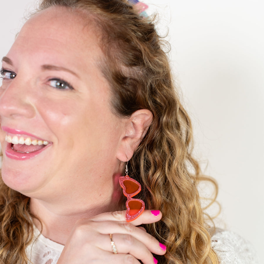 Heart-shaped earrings on a 'cheerful' branded card against a pink background