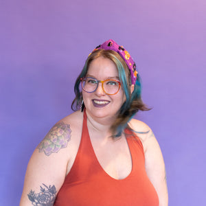 Person wearing an orange dress with colorful hair and glasses against a purple background