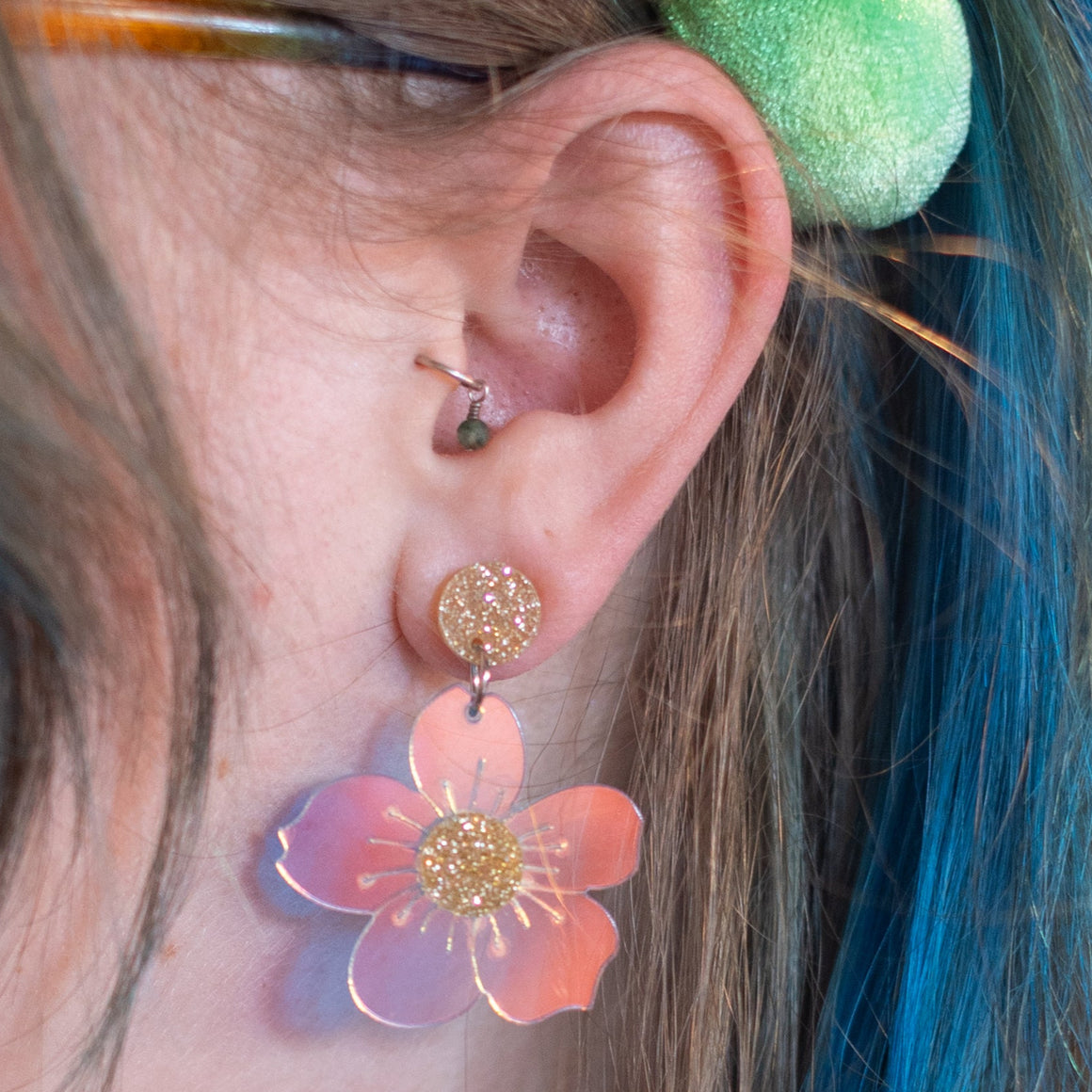 Person with colorful hair and headband, wearing glasses and floral earrings against a pink background