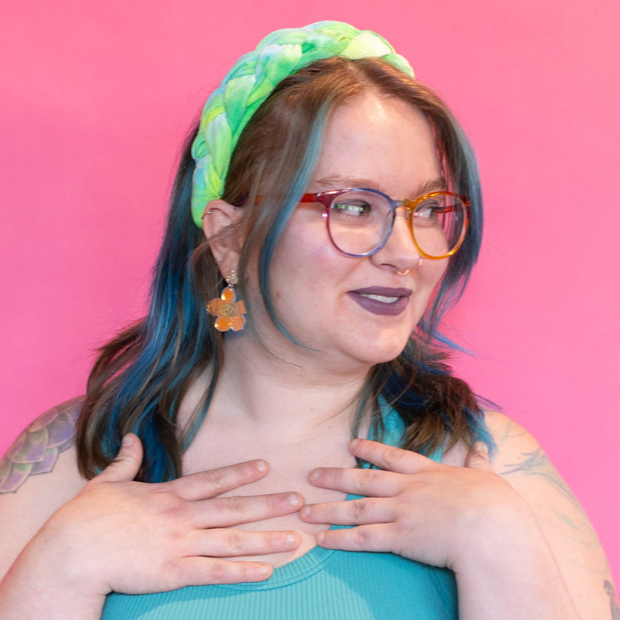 Person with colorful hair and headband, wearing glasses and floral earrings against a pink background