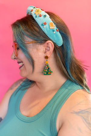 Person wearing a turquoise headband and earrings with colorful decorations against a pink background