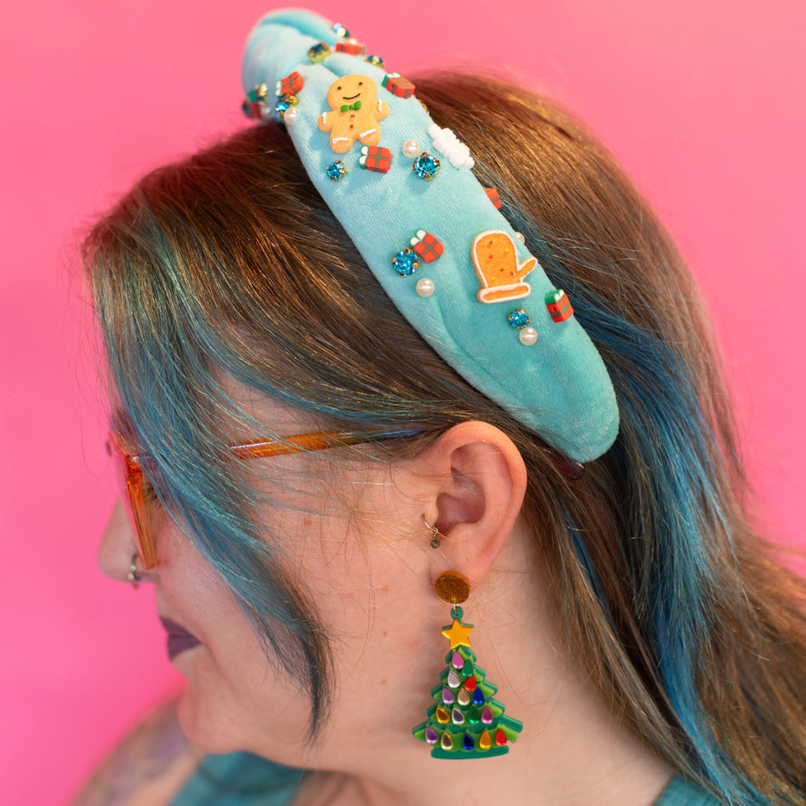 A light blue headband with a Christmas-themed design, featuring gingerbread men, snowflakes, and rhinestones, displayed against a holiday-themed background with a green and red color scheme and snowflake decorations.
