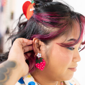 Person wearing colorful earrings with strawberry design, close-up of face and earrings.