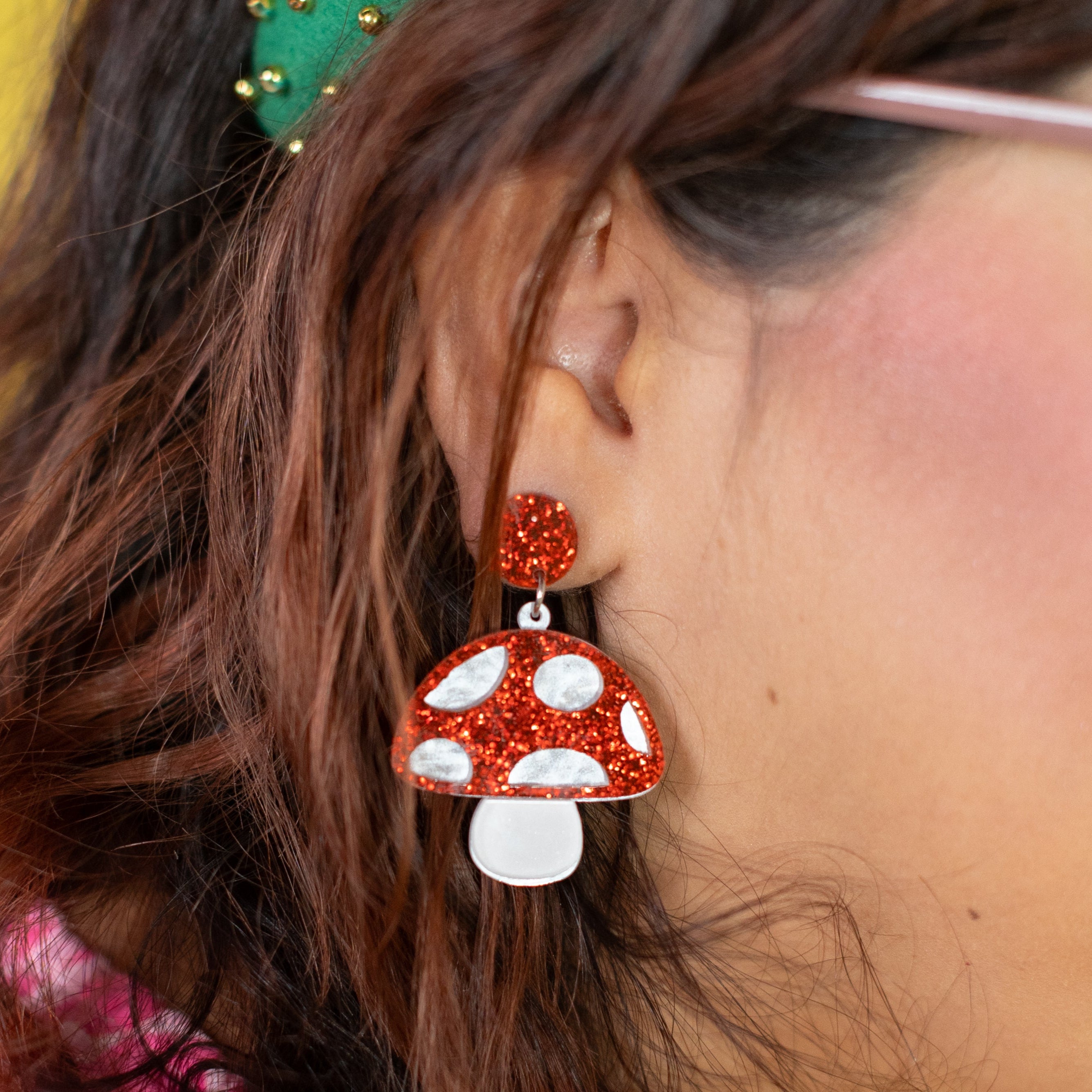 Close-up of a person wearing red and white mushroom-shaped earrings with a green headband against a yellow background.