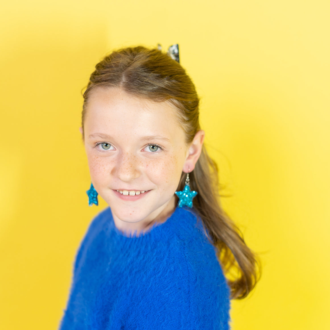 Young girl wearing a blue sweater and earrings against a yellow background