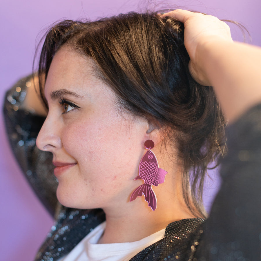 Pair of purple fish-shaped earrings on a 'chearful' branded card against a pink background