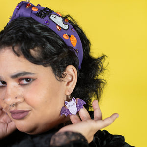 Person wearing a purple headband and matching earrings with cat designs against a yellow background