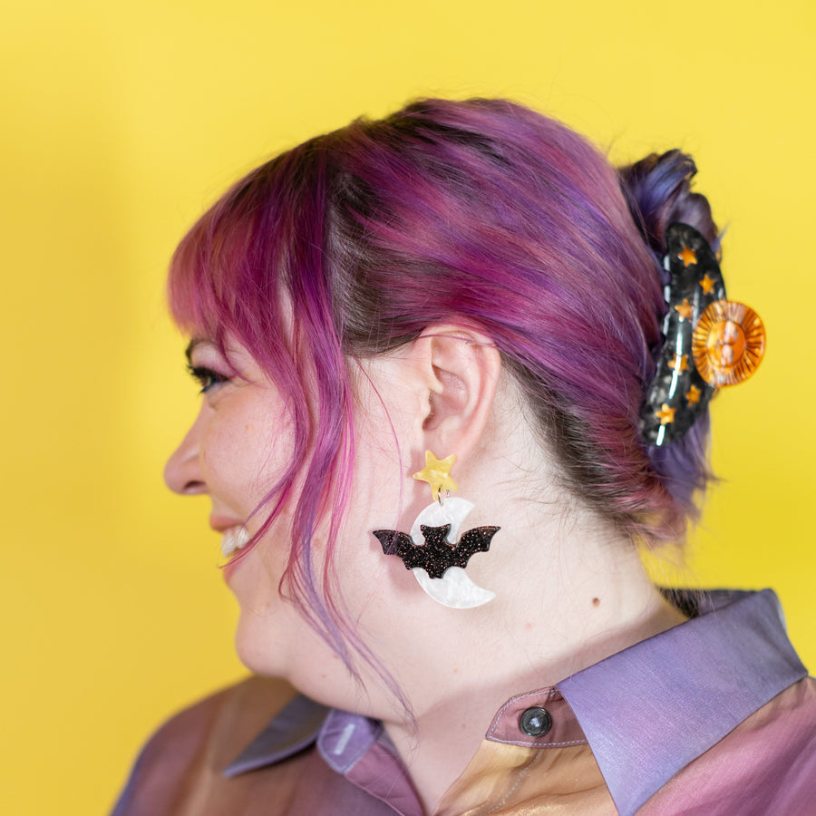 Pair of black bat-shaped earrings on a 'cheerful' branded card against a purple background