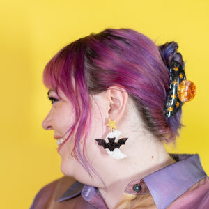 Person with purple hair wearing bat-shaped earrings against a yellow background