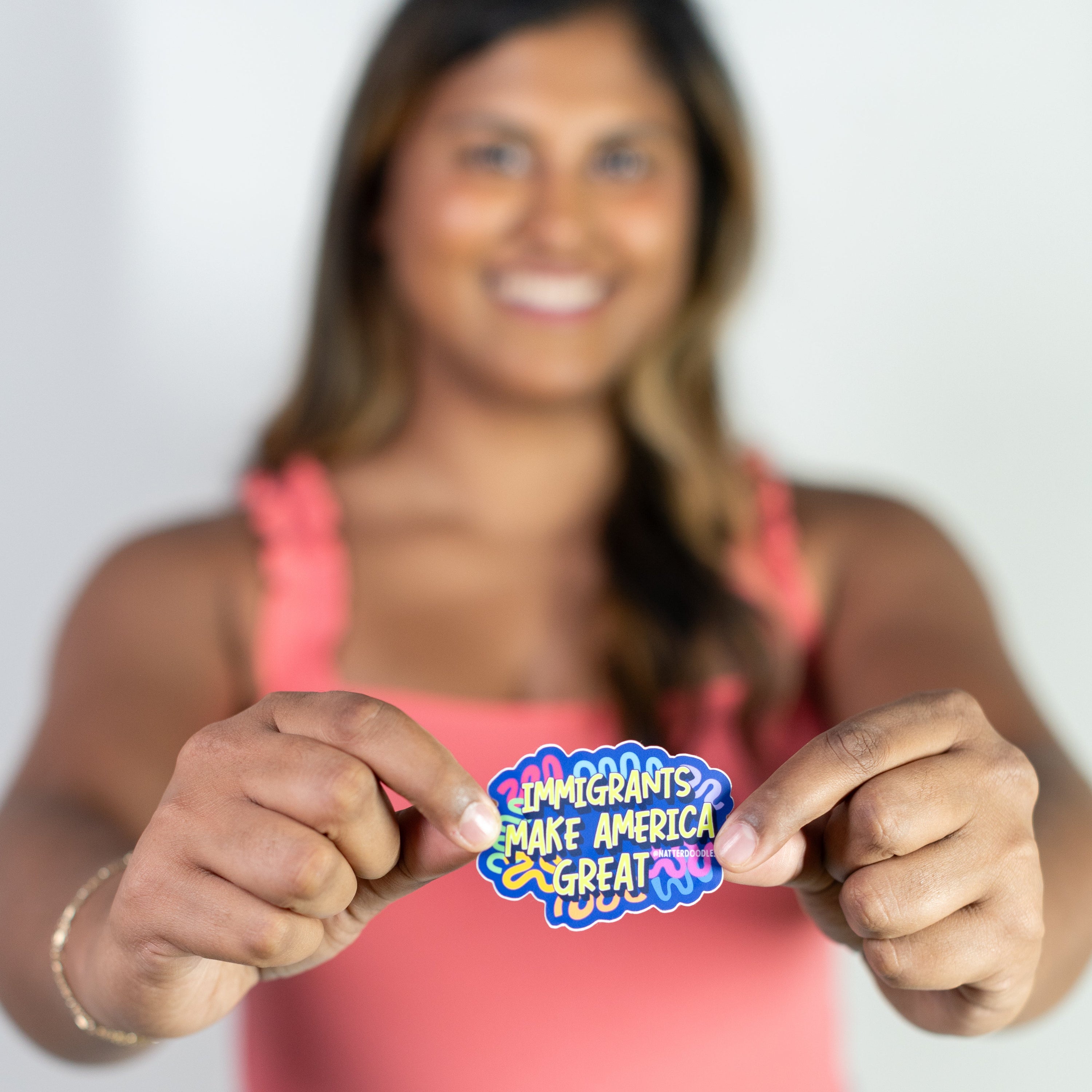 Woman holding a colorful sticker with text, wearing a pink top and blue jeans.
