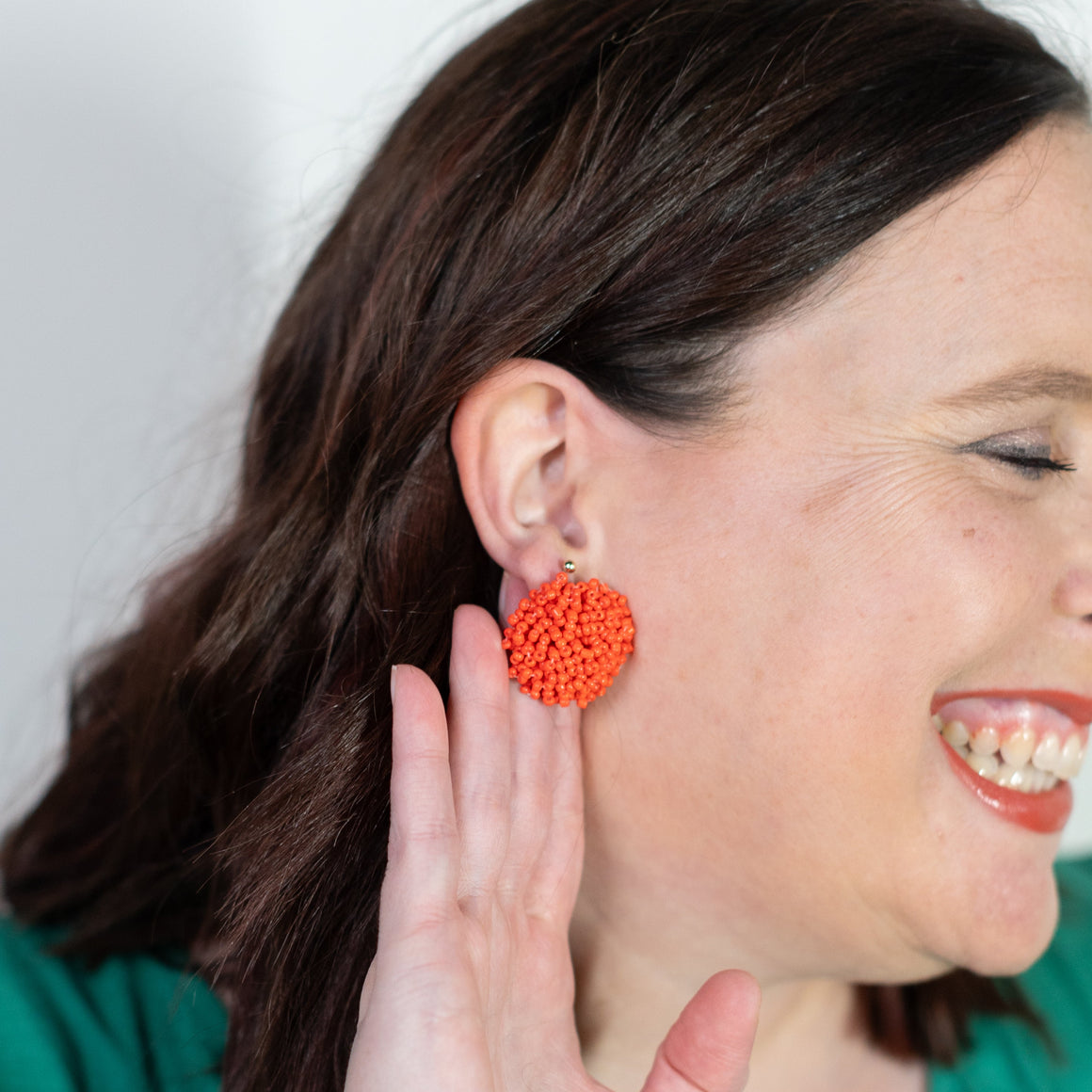 Woman wearing orange pom-pom earrings, smiling and touching her ear.