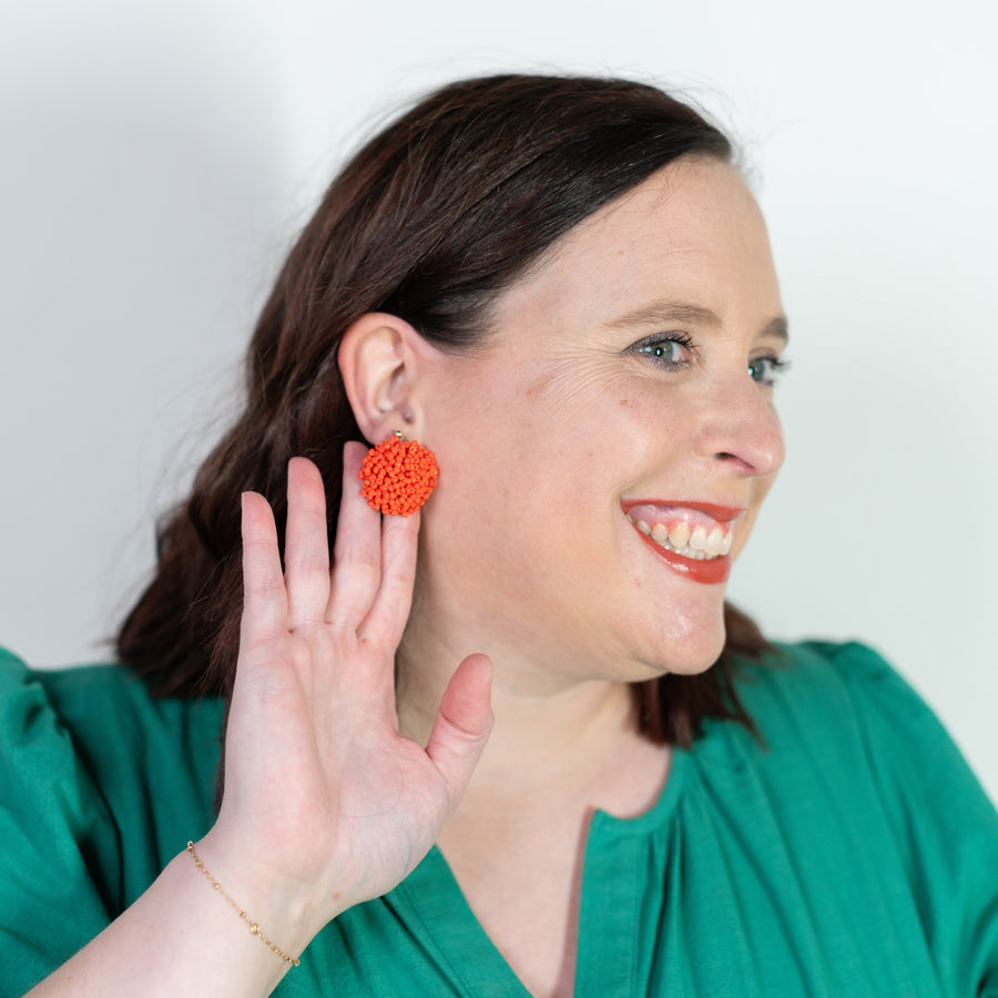 Woman wearing orange pom-pom earrings, smiling and touching her ear.