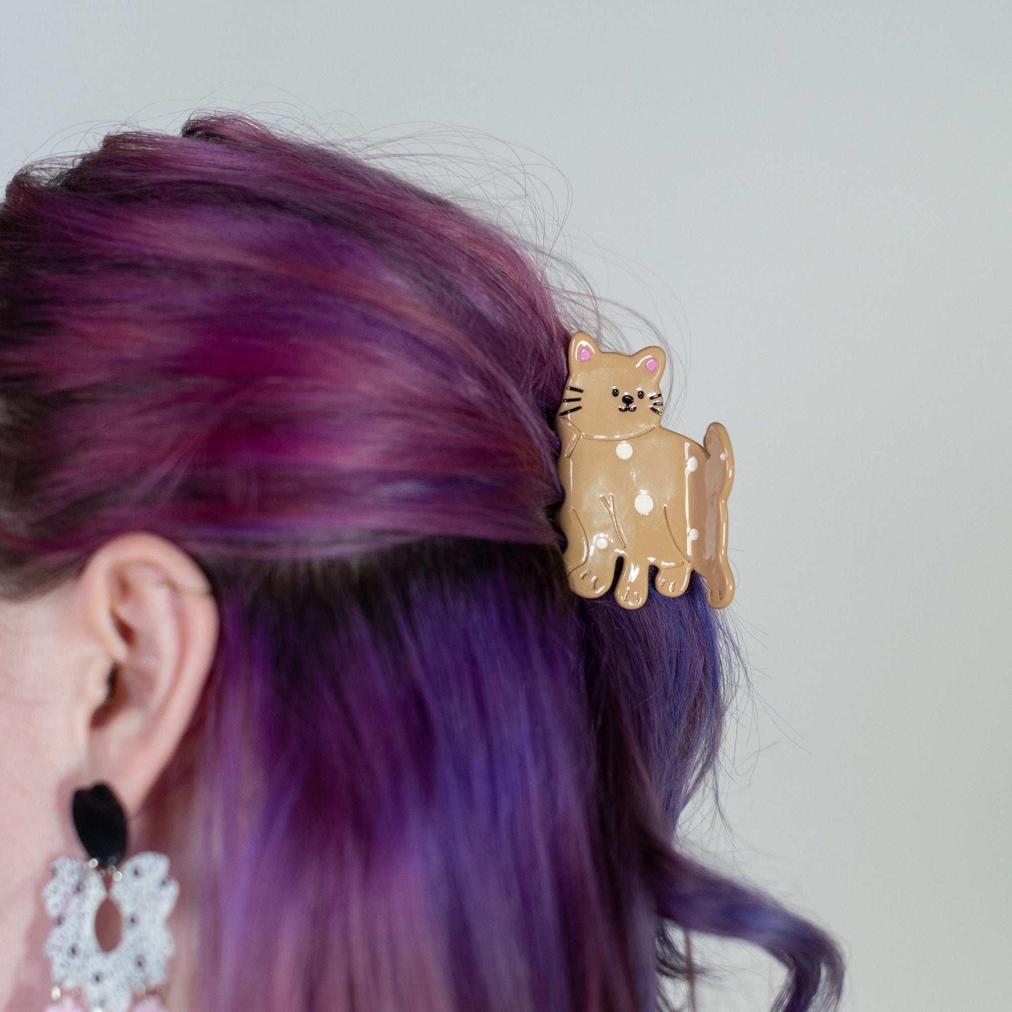 Person with purple hair wearing a gold cat-shaped hair clip against a plain background