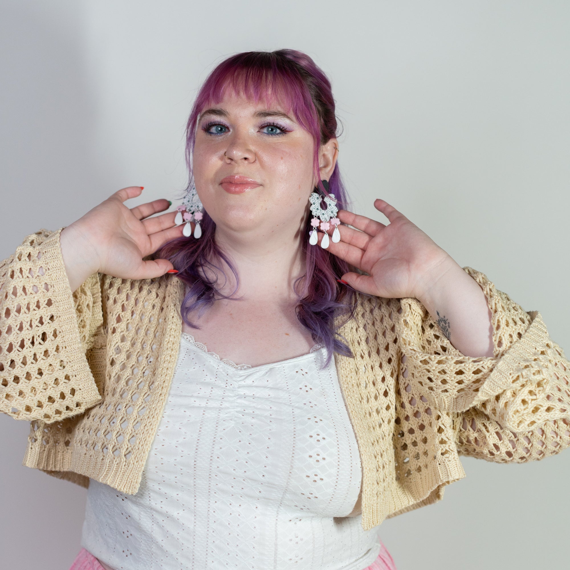 Person wearing a beige crochet cardigan, white top, and pink skirt with purple hair against a plain background