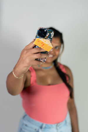 Person holding a phone with a colorful case and a piece of cheese, wearing a pink top and blue jeans.