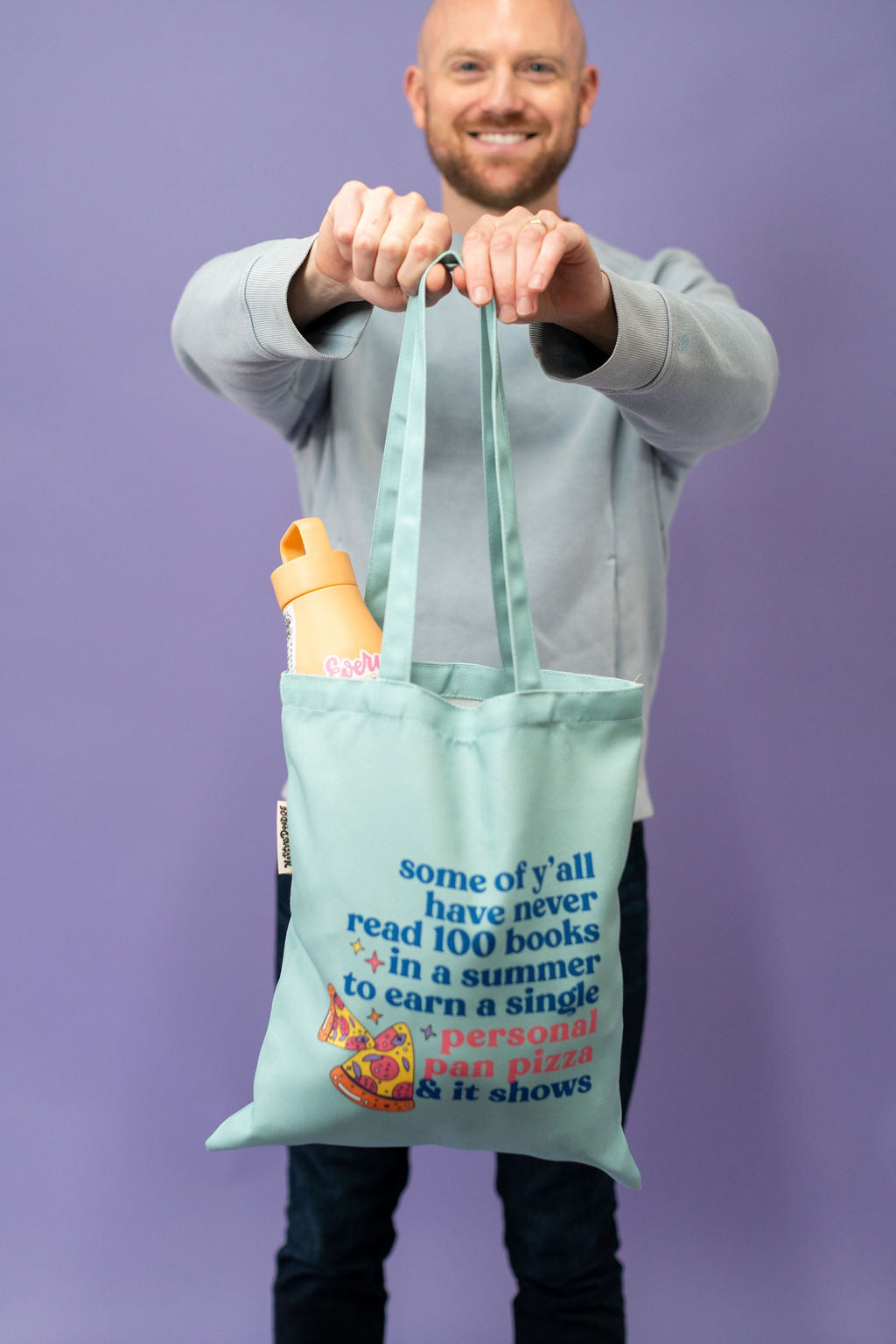 Man holding a light blue tote bag with text and a bottle against a purple background