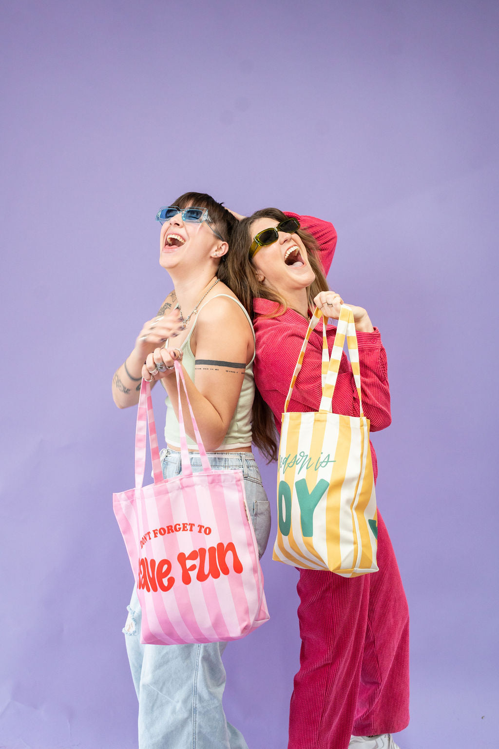 Two people holding colorful tote bags against a purple background
