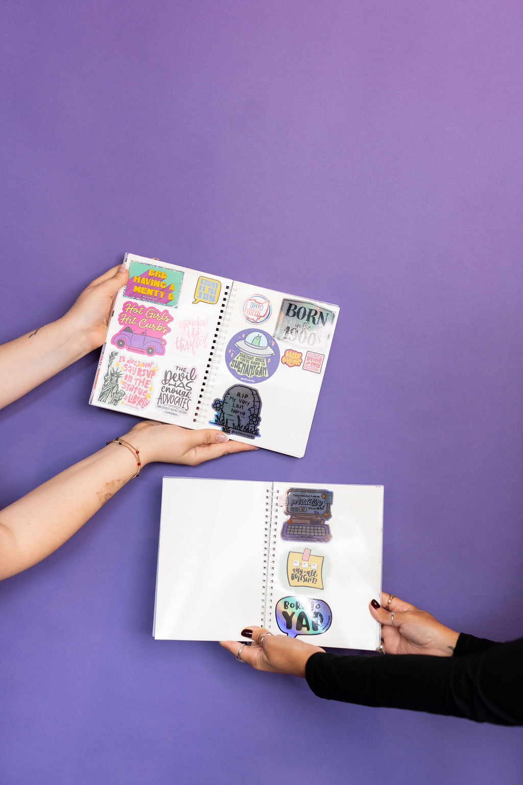 Two people holding open sticker books with colorful stickers on a purple background