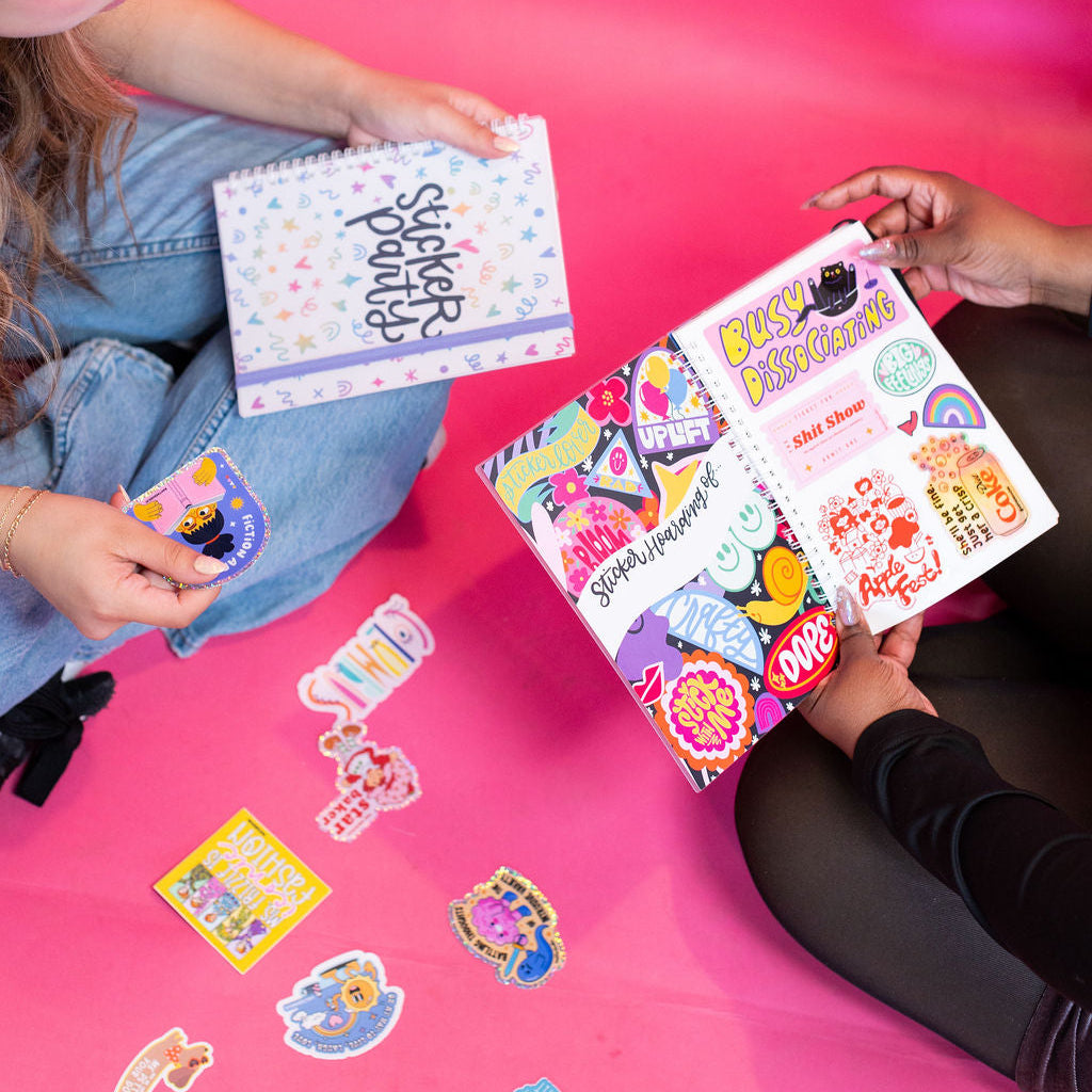 Two people sitting on a pink surface with sticker books and stickers.