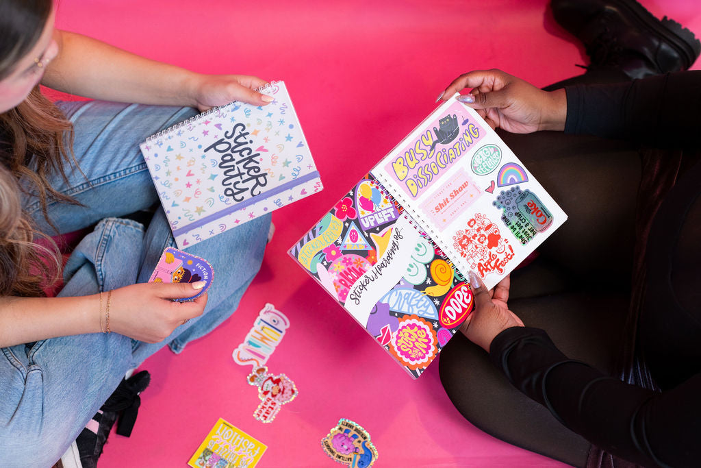 Two people sitting on a pink floor with sticker books and stickers.