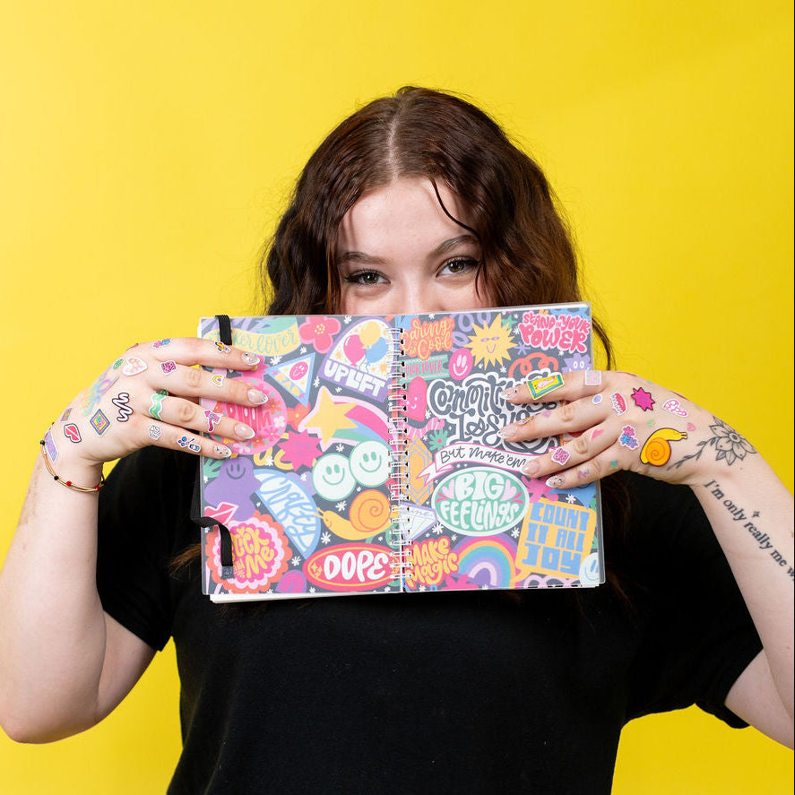 Person holding a colorful patterned sticker book in front of their face against a yellow background