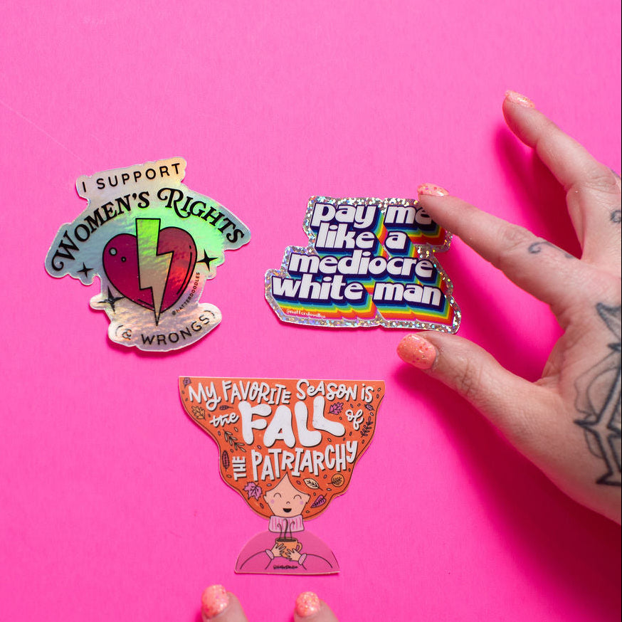 Three protest-themed stickers on a pink background with hands holding them.