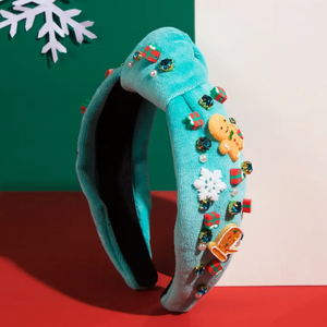 A light blue headband with a Christmas-themed design, featuring gingerbread men, snowflakes, and rhinestones, displayed against a holiday-themed background with a green and red color scheme and snowflake decorations.