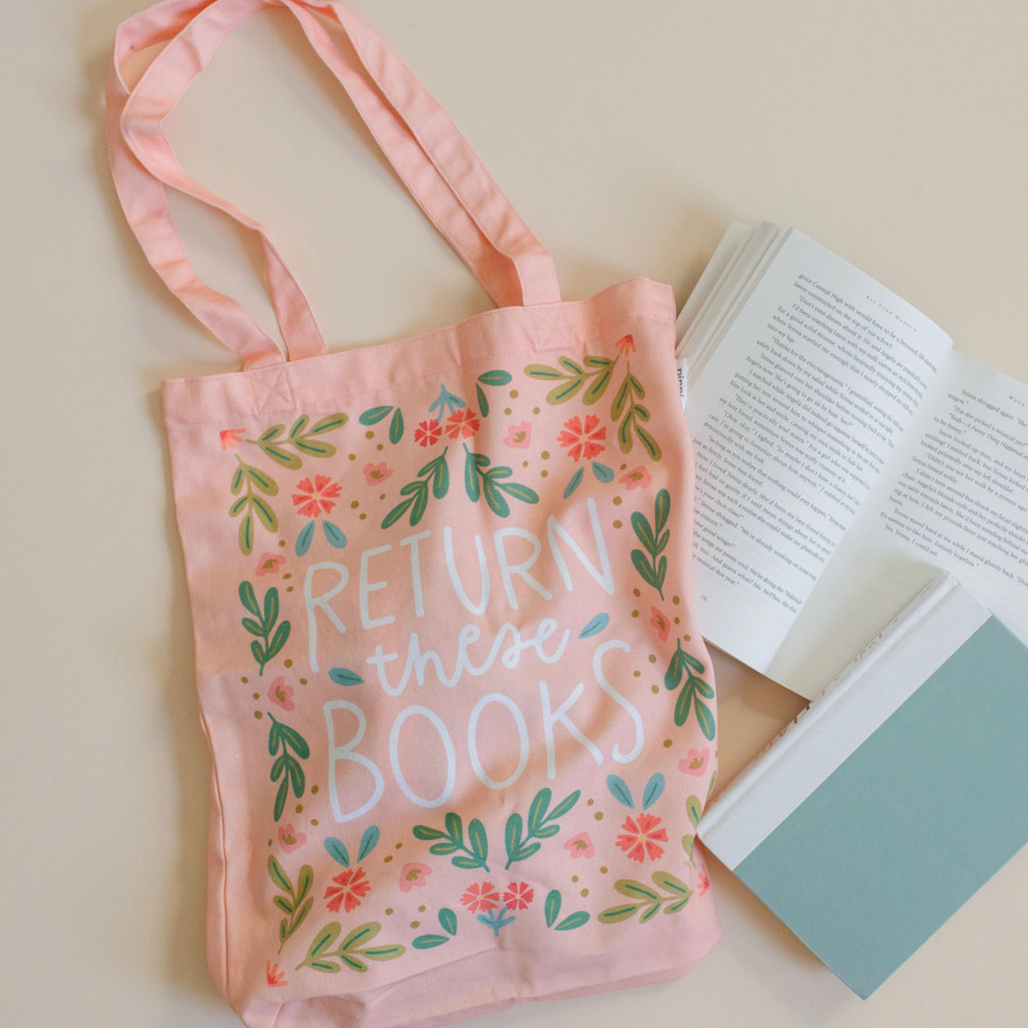 Pink tote bag with 'RETURN these BOOKS' text and floral designs on a light surface with books.