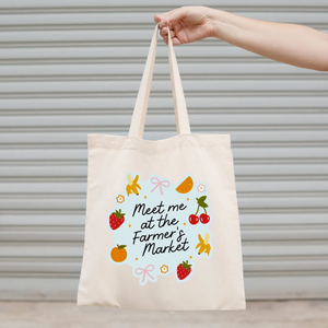 Tote bag with 'Meet me at the Farmer's Market' text and graphics held by a hand against a neutral background