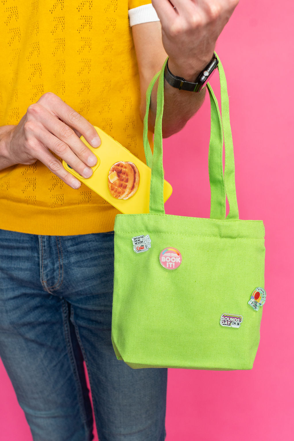 Person holding a green tote bag with colorful pins against a pink background