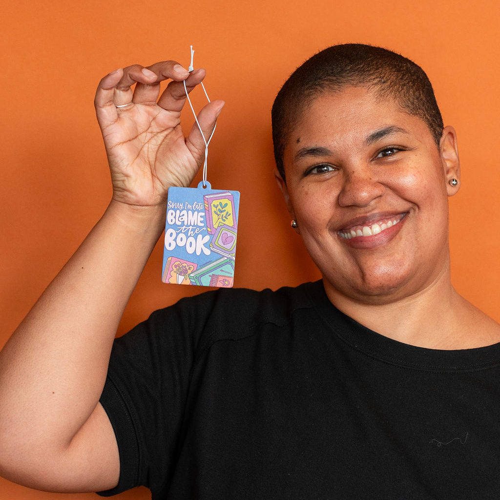 Person holding a car air freshener with a colorful label against an orange background