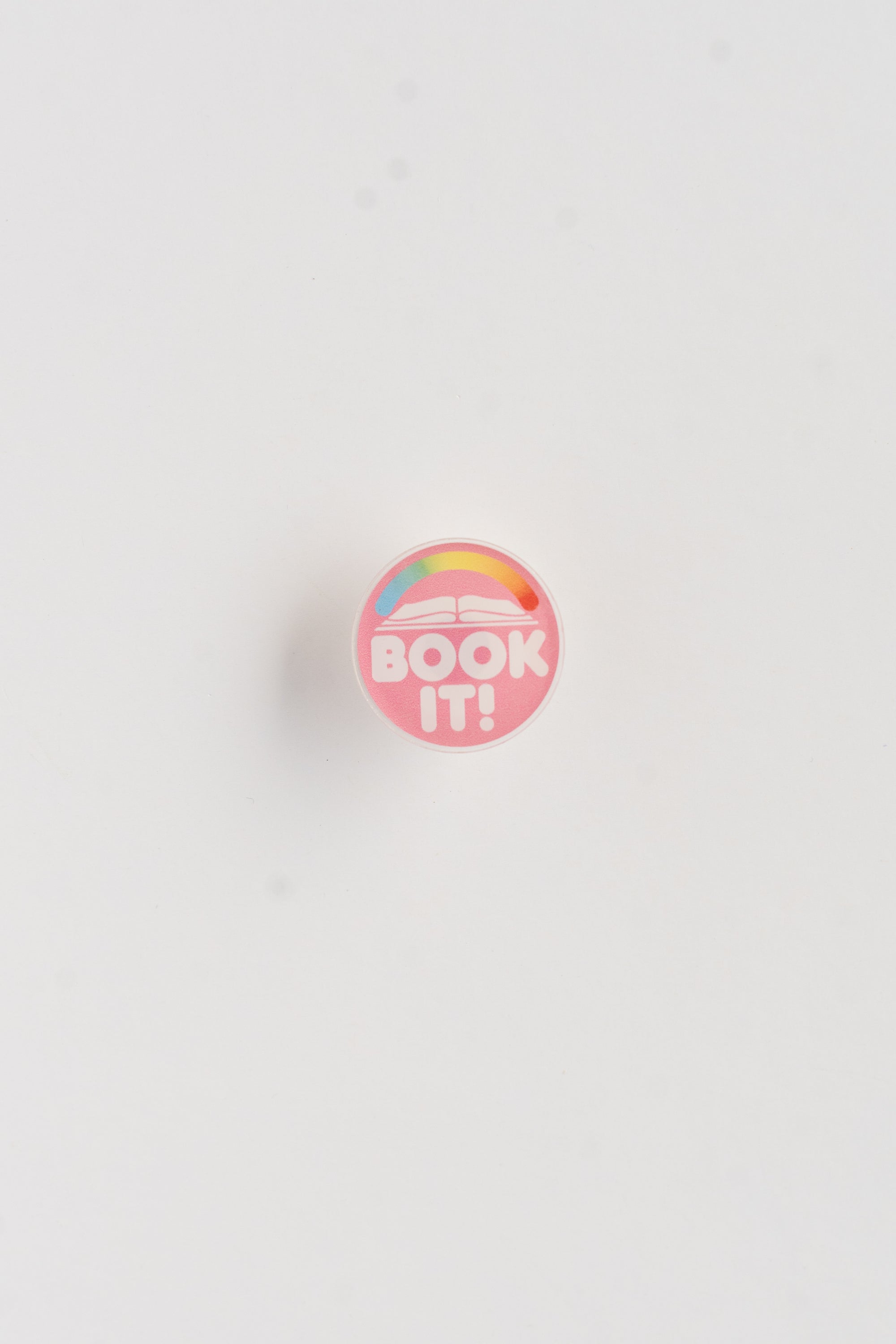Round pink button with 'BOOK IT!' text and rainbow design on a white background